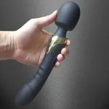 Black Dual Head Vibrator held in one hand, showing the rounded head, curved lower end and gold-tone control panel